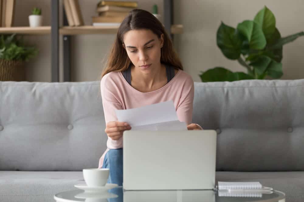 A woman sitting on a couch with a rejection letter in her hand and a coffee cup and laptop on the table in front of her.